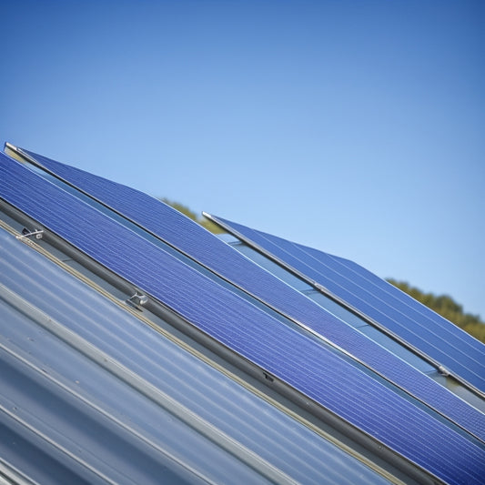 A close-up of a silver aluminum solar panel rack, mounted on a sloping roof, with a subtle shadow on the roof's surface, and a faint blue sky in the background.